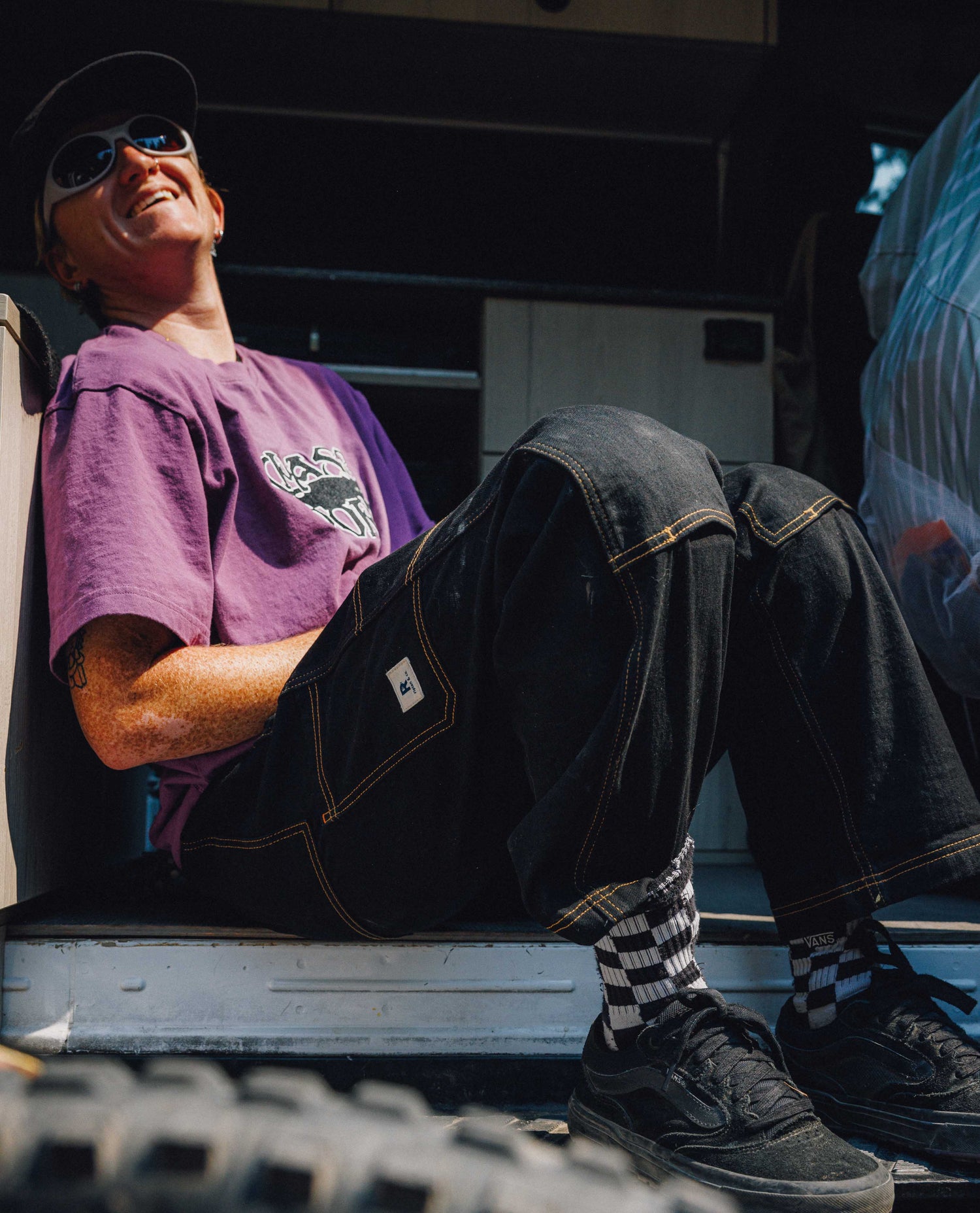 Woman sitting wearing black pants with yellow stitching, a purple shirt, and gray sunglasses.