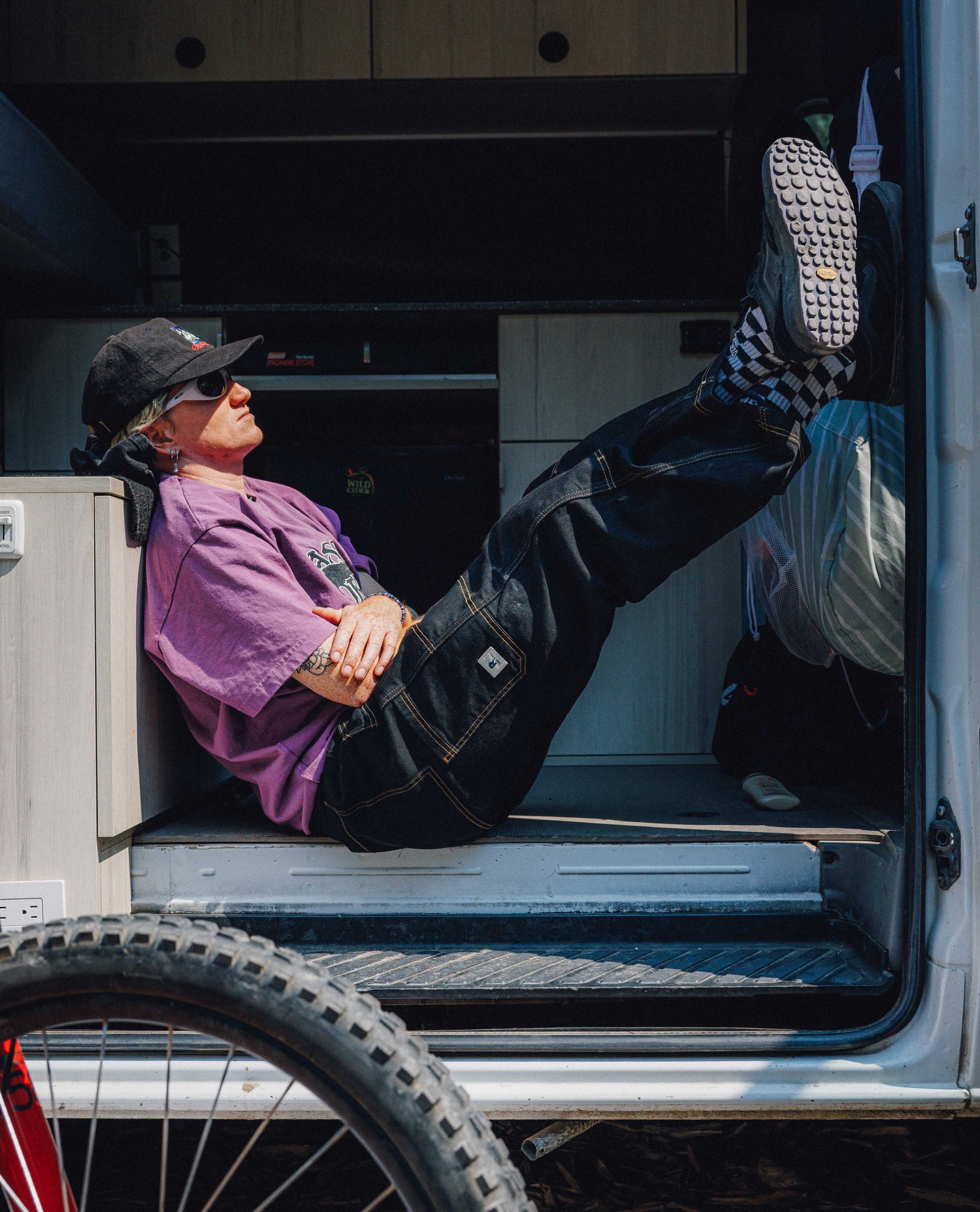 Woman sitting with legs up wearing checkered socks, purple shirt, black hat, gray sunglasses, and black pants with yellow stitching