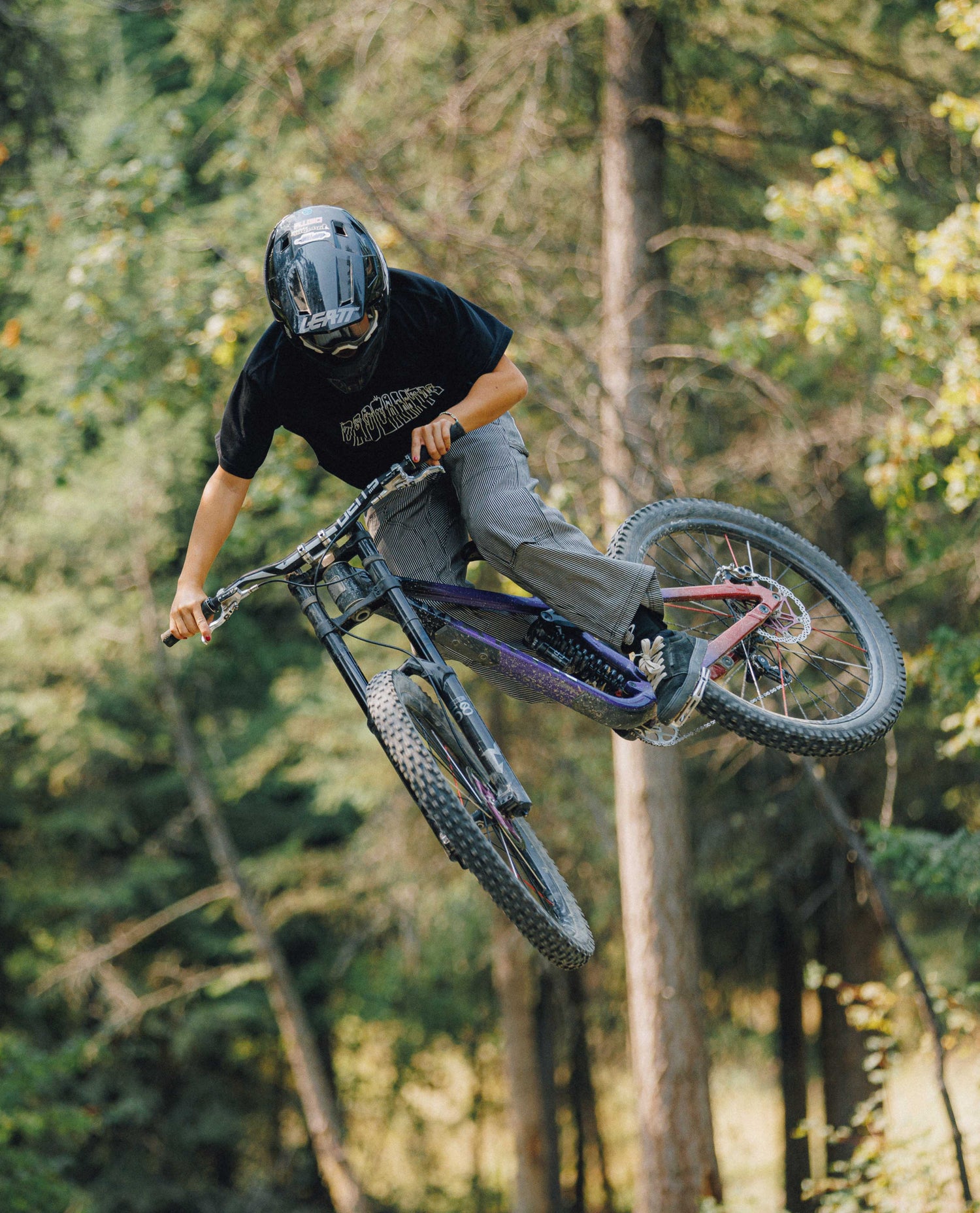 Woman on a bike in midair, in front of trees. She is wearing a black shirt and black and white striped pants.