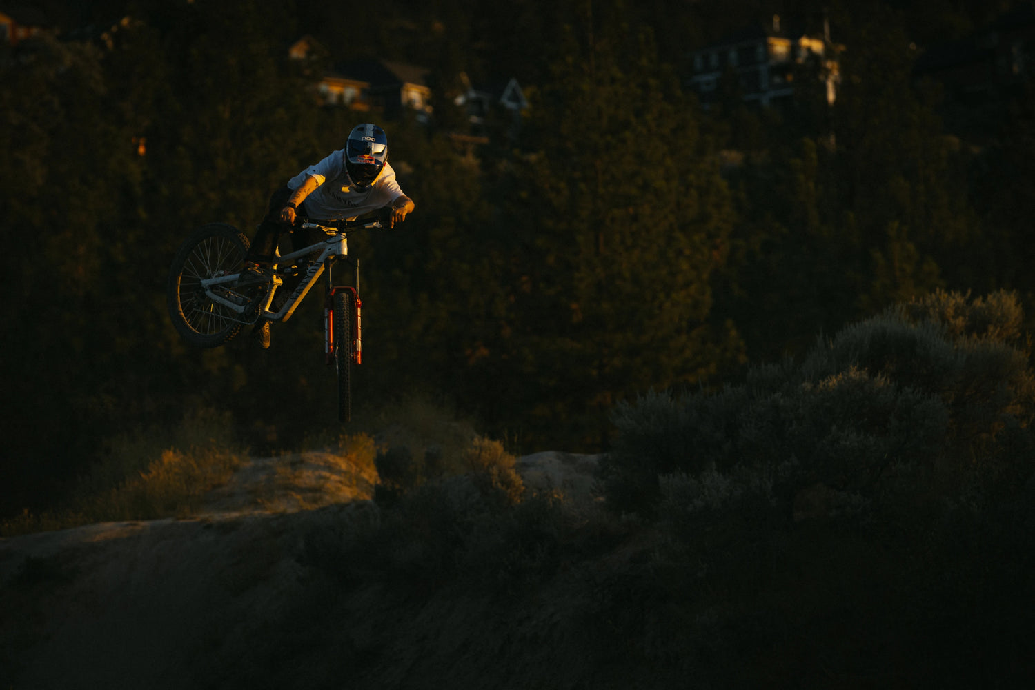 Person on a mountain bike in mid-air during a jump at dusk with trees and lights in the background.