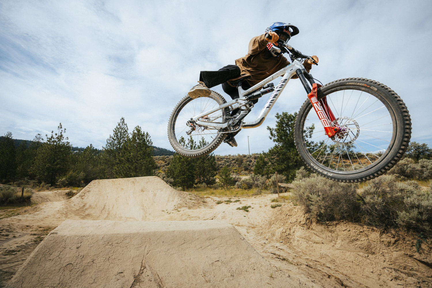 Biker performing a jump on a dirt bike track with trees and sky in the background