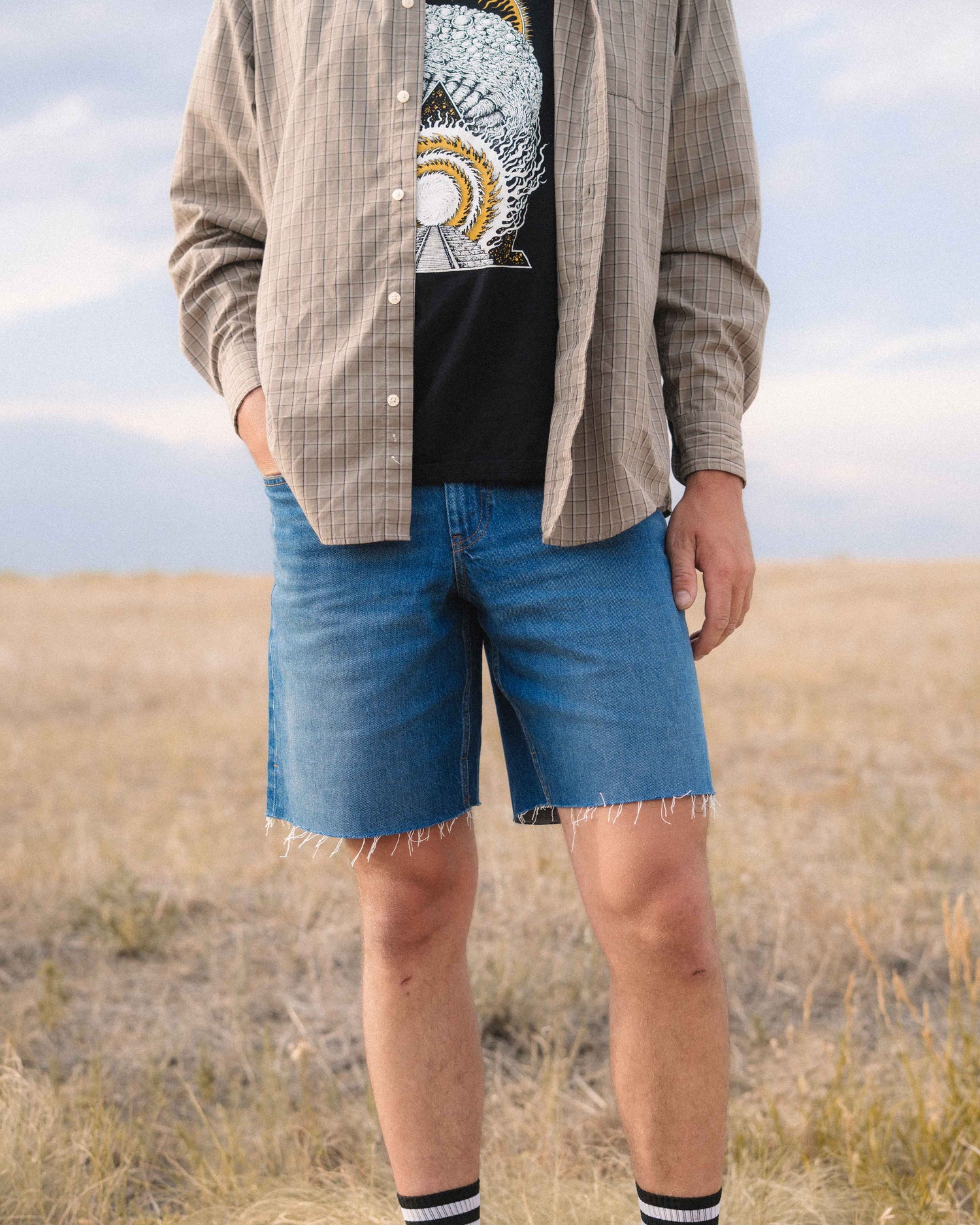 A person standing outdoors in a grassy field wearing medium-wash blue denim shorts with a frayed hem, a black graphic T-shirt, and an open beige plaid button-up shirt.