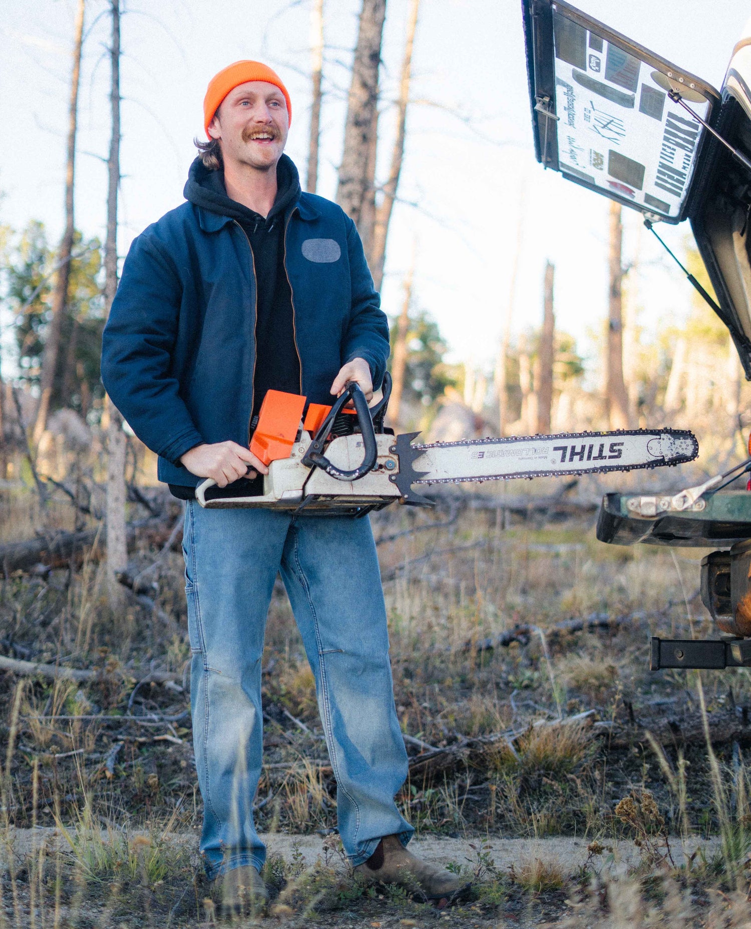 Man holding a chainsaw in a forest setting with a vehicle in the background