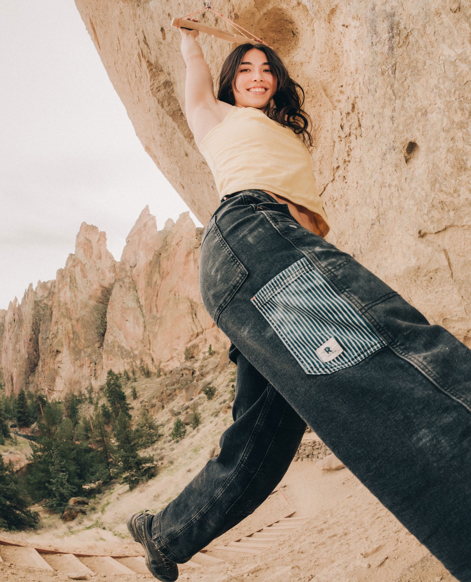 Girl in front of rocks outside, wearing blue pants with striped pocket and yellow shirt. 