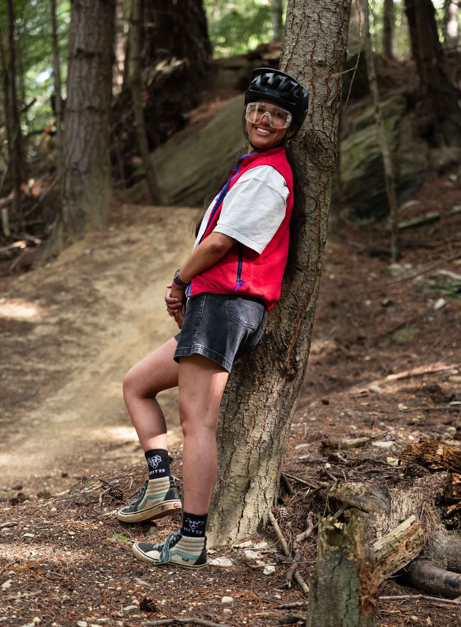 A woman wearing a red vest, white shirt, black shorts, and helmet leaning against a tree