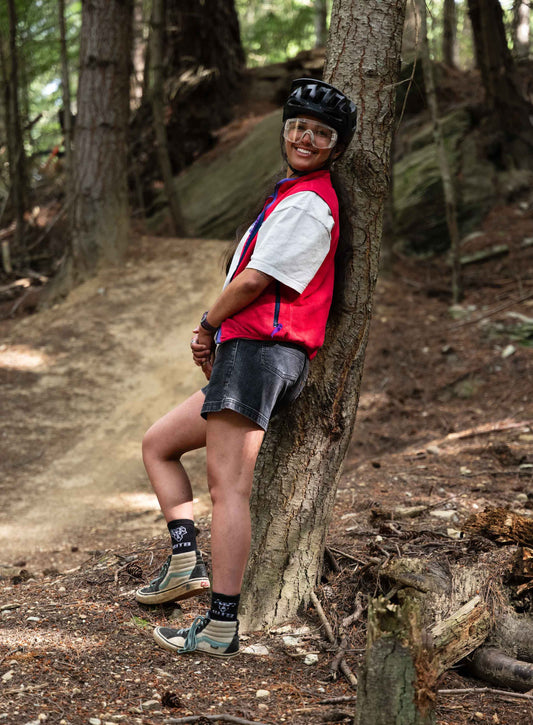 A woman wearing a red vest, white shirt, black shorts, and helmet leaning against a tree
