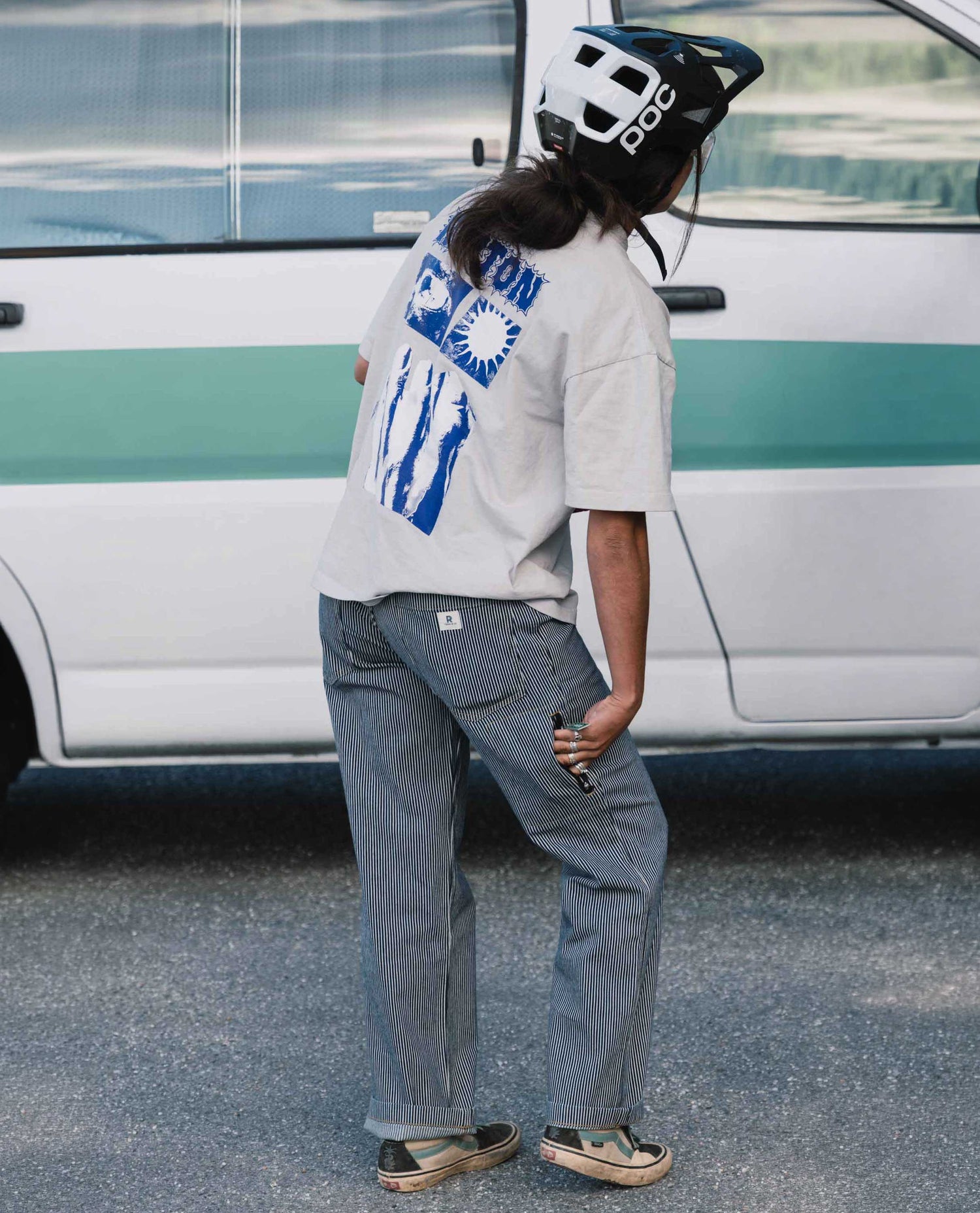 Person wearing a white t-shirt with blue graphics and gray pants standing in front of a van.
