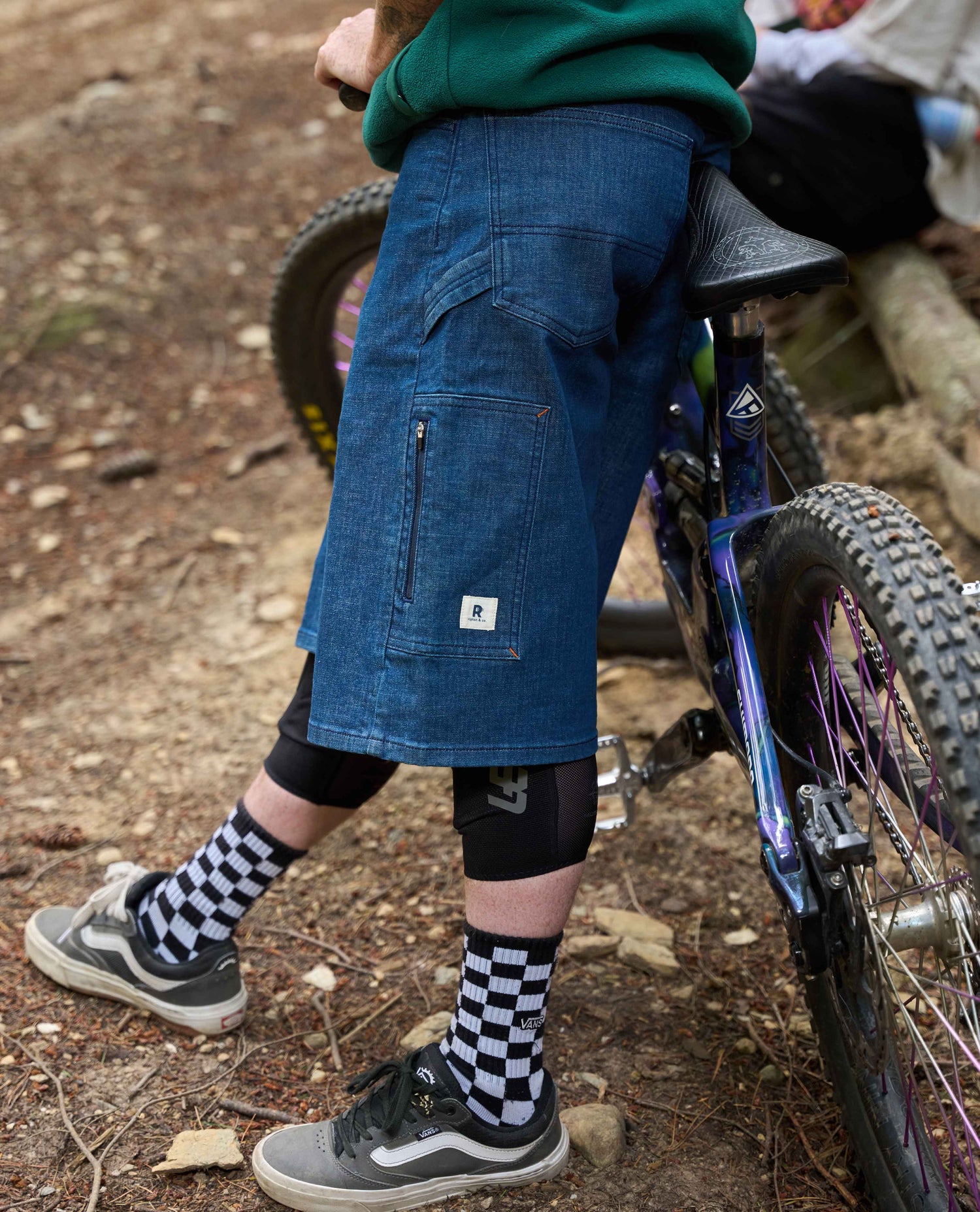 Person wearing blue shorts and checkered socks standing next to a bicycle on a dirt path.