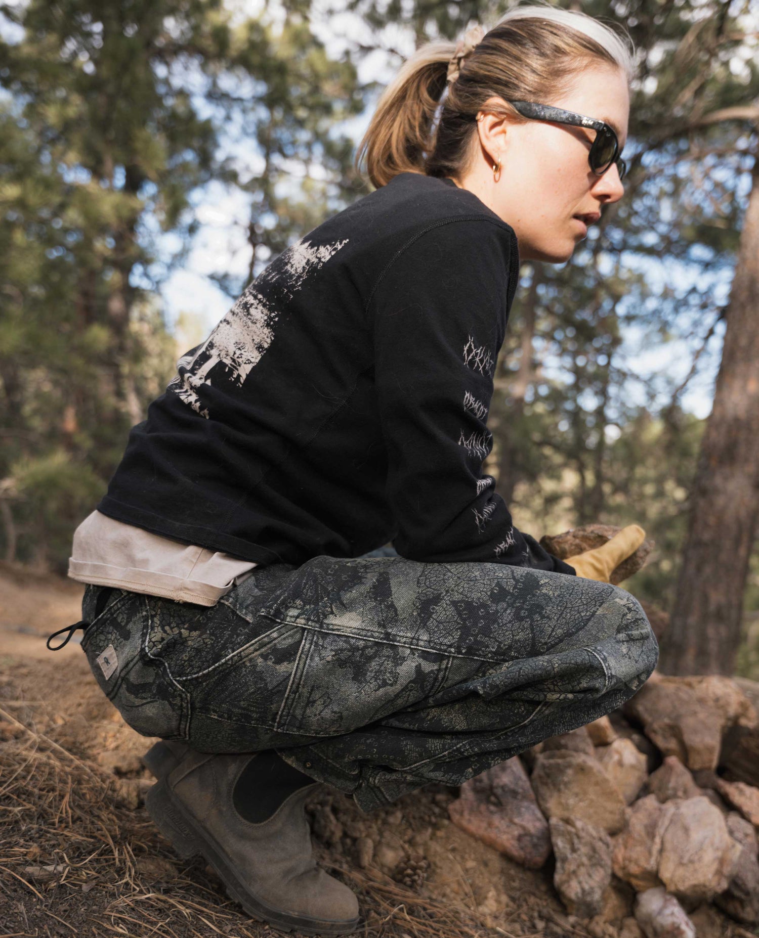Woman crouching and wearing bark leaf camouflage pants outside. 