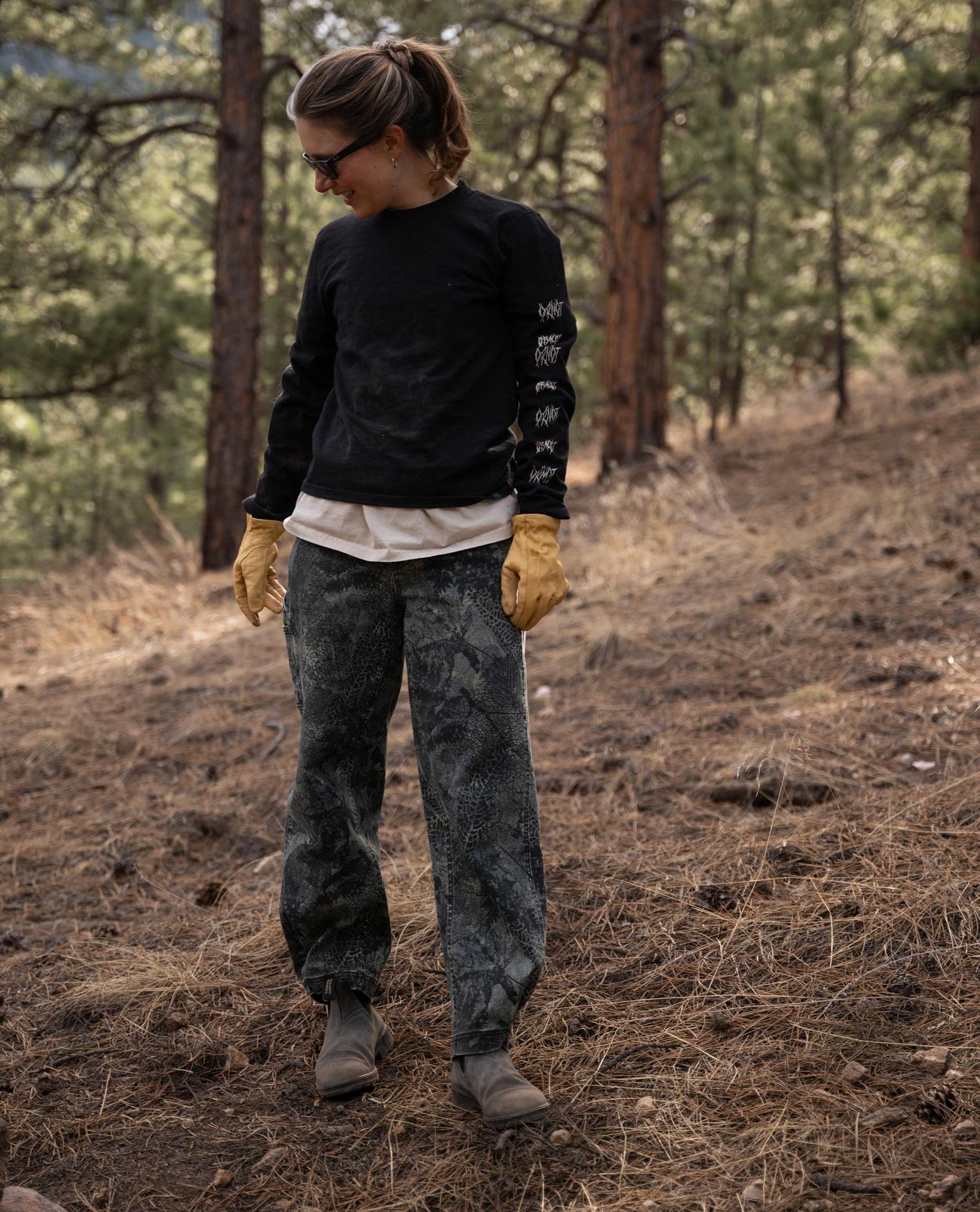 Woman wearing camouflage leaf pants and black shirt, standing outside. 