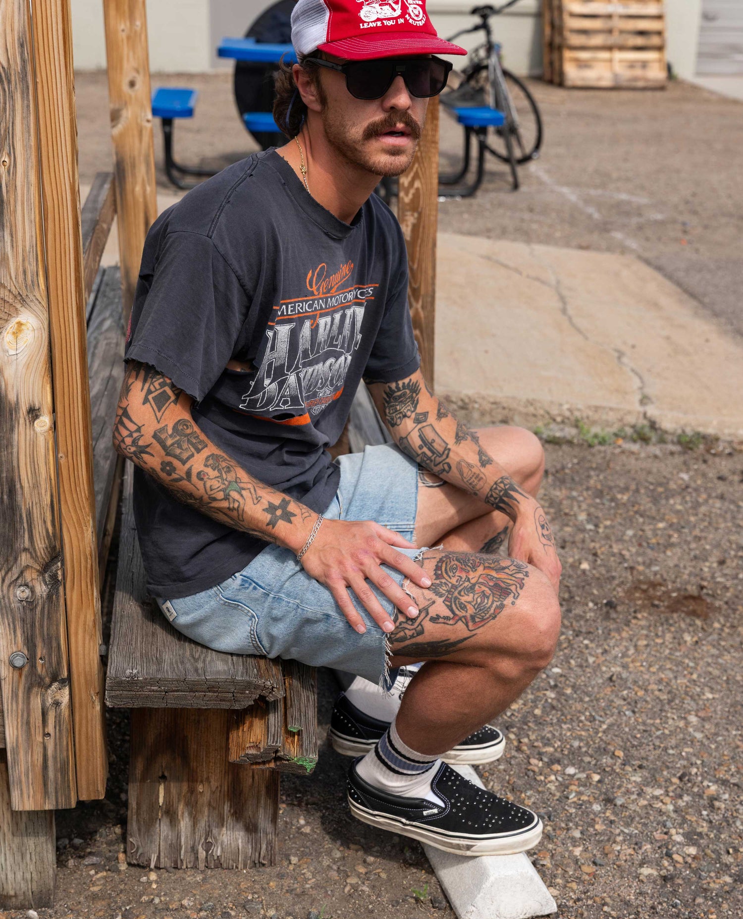 Man with tattoos wearing a red cap and sunglasses, sitting on a wooden bench outdoors.