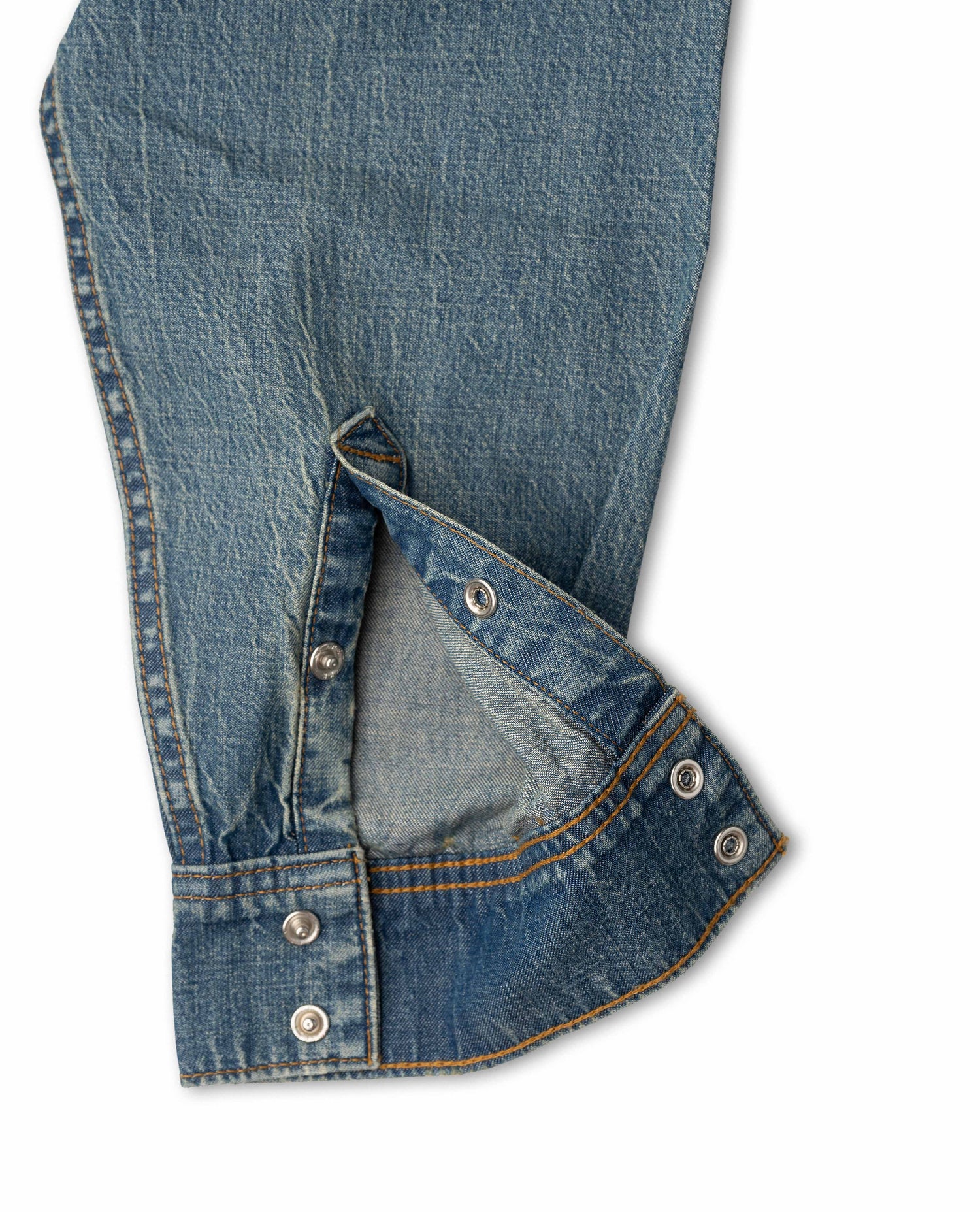 Close-up of a blue denim garment, showing the fabric texture, stitching, and silver metal snap buttons on a white background.