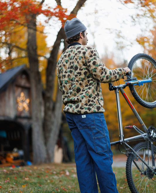 Person in camouflage jacket and blue pants standing next to a bicycle with autumn trees in the background
