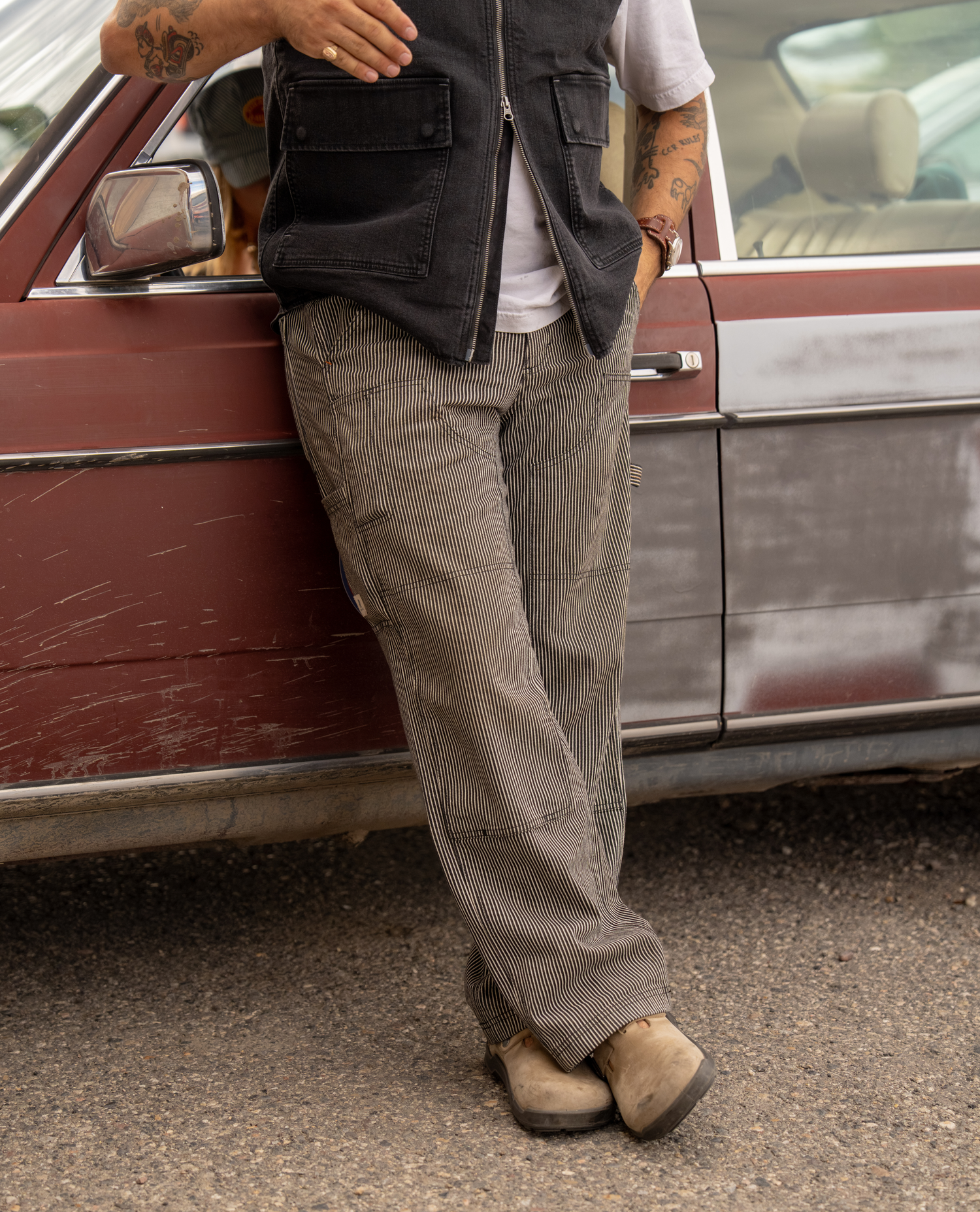 A person wearing black and white striped pants, a black zip vest, and tan boots leans against a vintage maroon car outdoors.