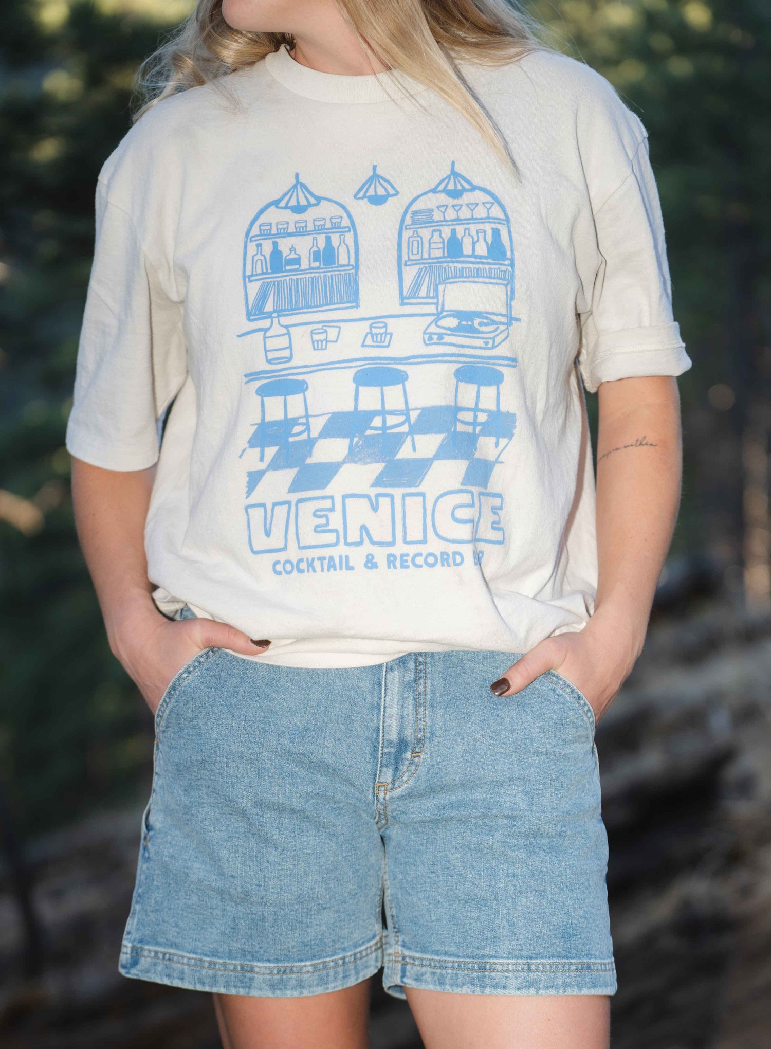 Close up of woman wearing blue shorts and white shirt outside, with hands in pockets 