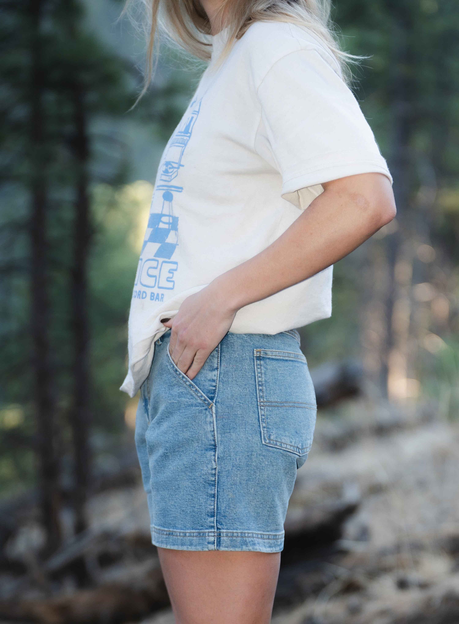 View of woman from the side wearing white shirt, blue shorts, standing outside with hands in pockets 