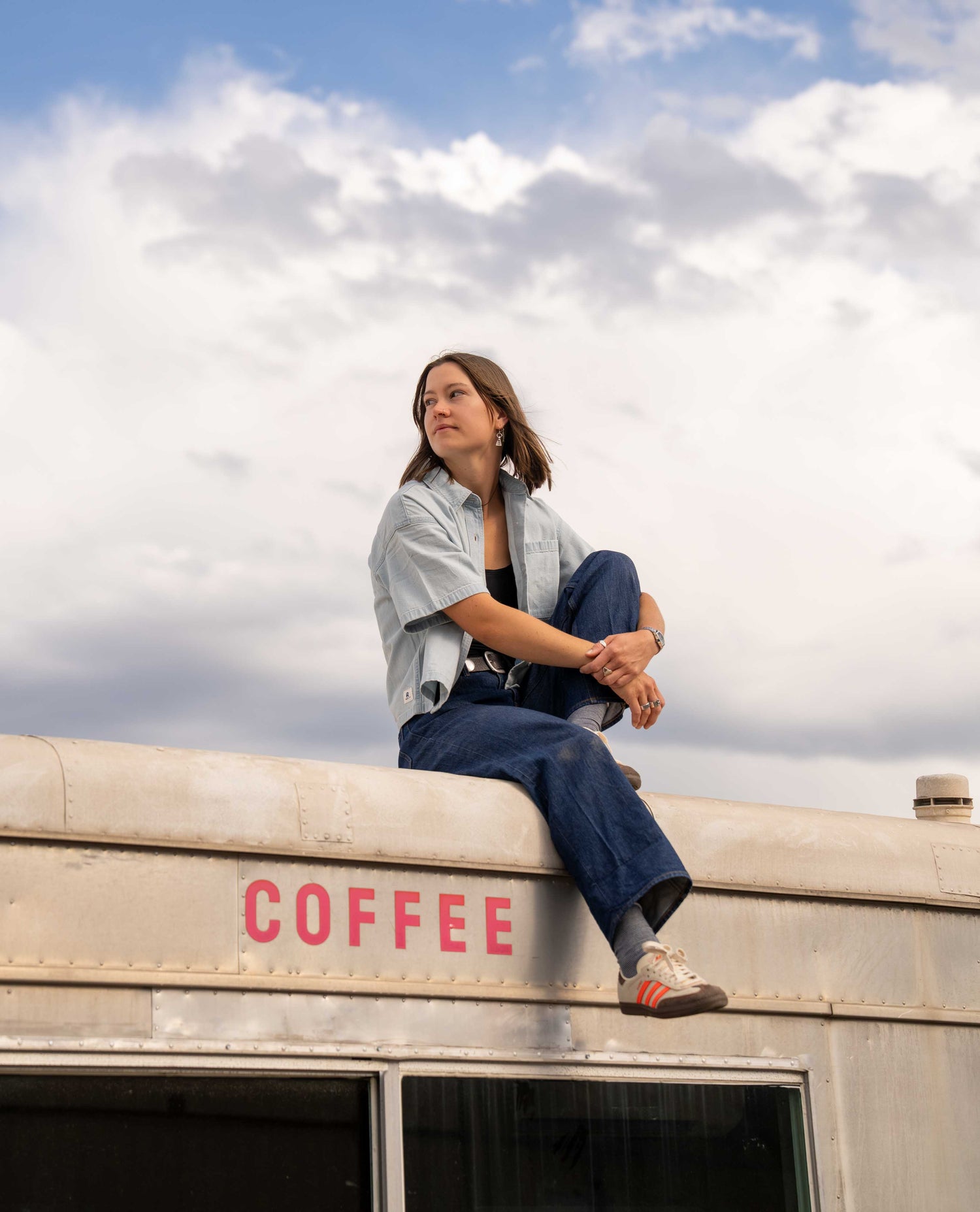 A woman sits on the roof of a vintage-style bus under a cloudy sky. She is wearing a light blue denim shirt, dark blue jeans, and white sneakers.