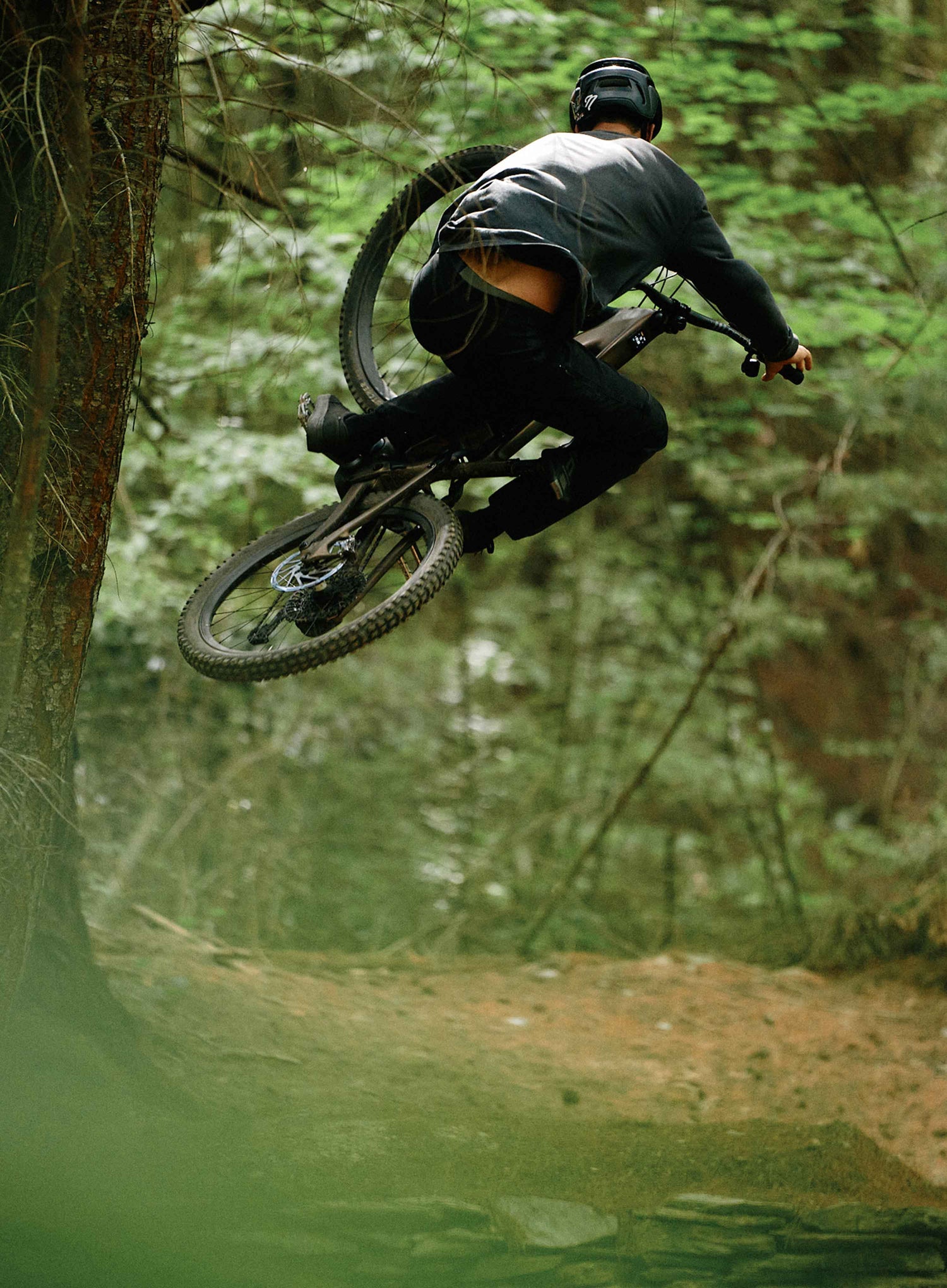 Person on a mountain bike in mid-air over a dirt trail with trees in the background