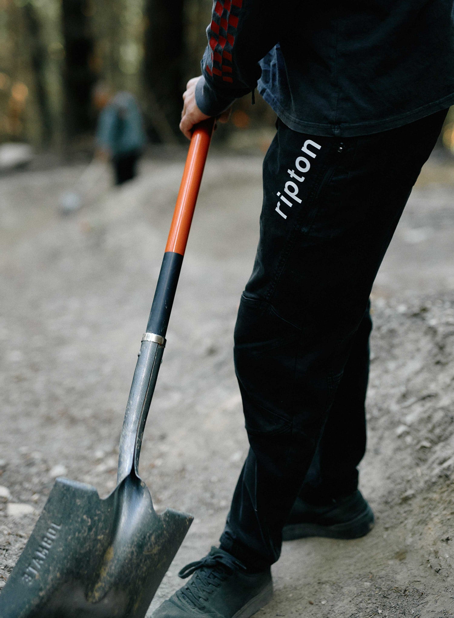 Person holding a shovel with 'ripton' branding on pants, blurred background
