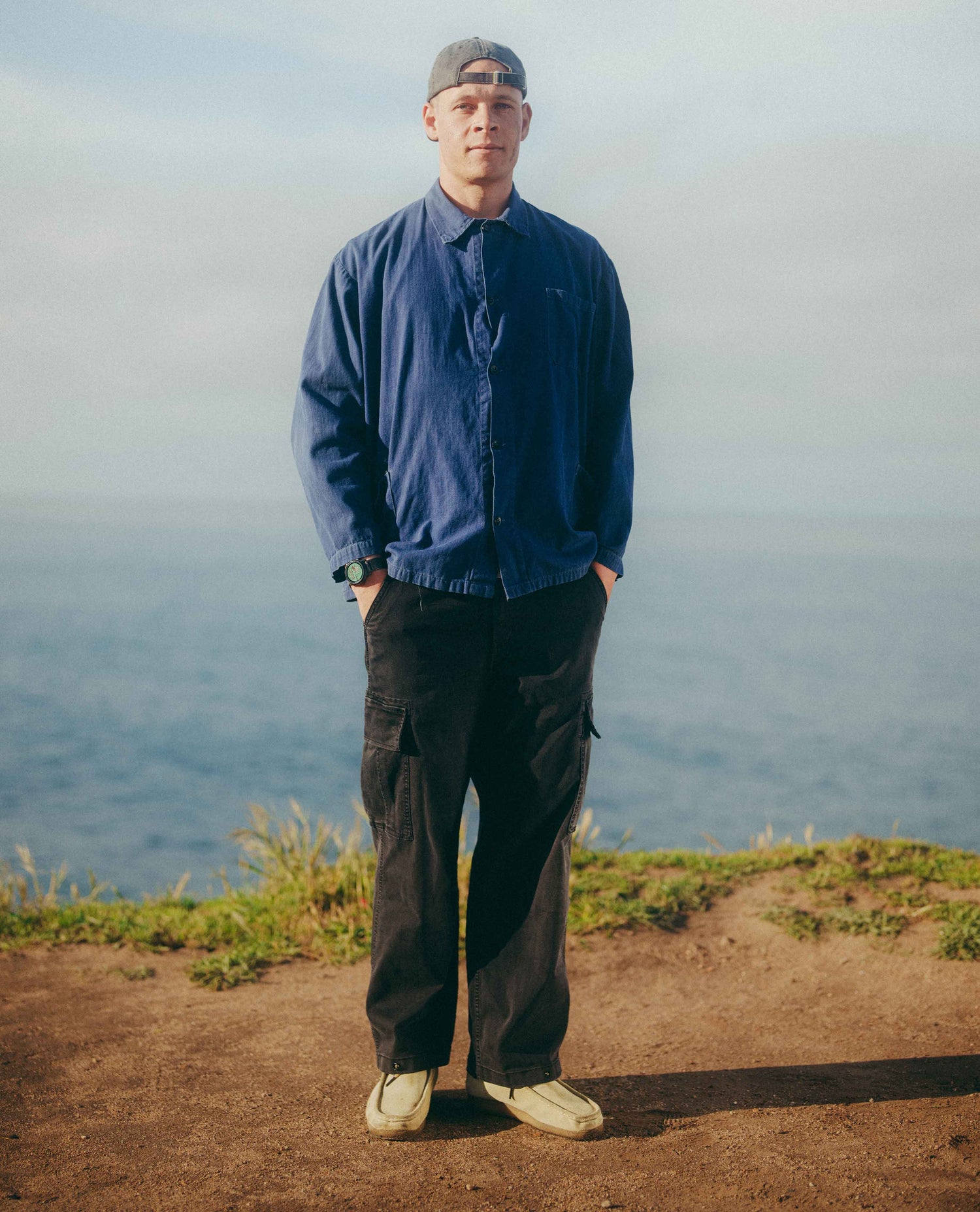 A man standing on a cliffside path with the ocean in the background, wearing a navy blue button-up shirt, dark cargo pants, a gray cap, and off-white sneakers.