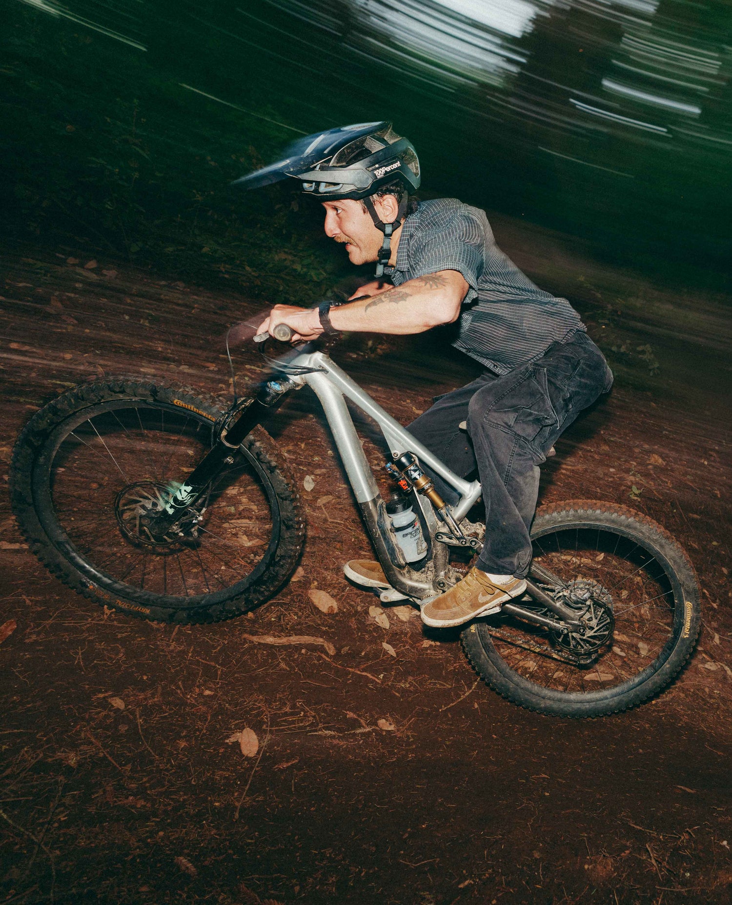 A mountain biker in motion on a dirt trail, wearing a dark helmet, gray shirt, dark pants, and brown shoes, with a blurred dark forest background.