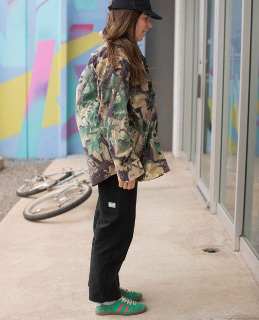 Person wearing a camouflage jacket and black pants standing on a sidewalk with a colorful mural in the background.