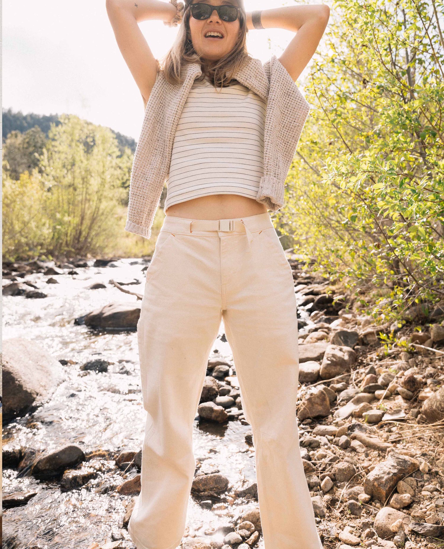 Person standing by a river wearing a striped shirt, vest, and beige pants with sunglasses.
