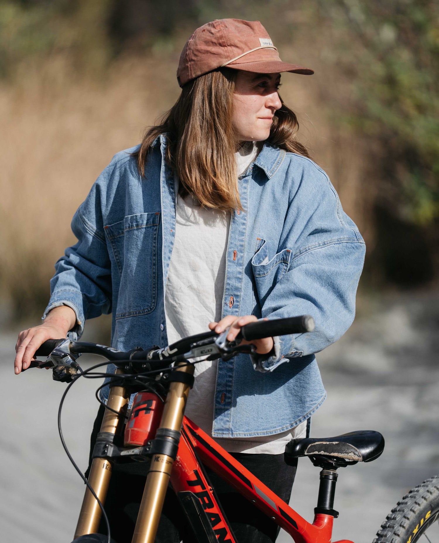 Person wearing a denim jacket and brown cap standing next to a red bicycle outdoors.