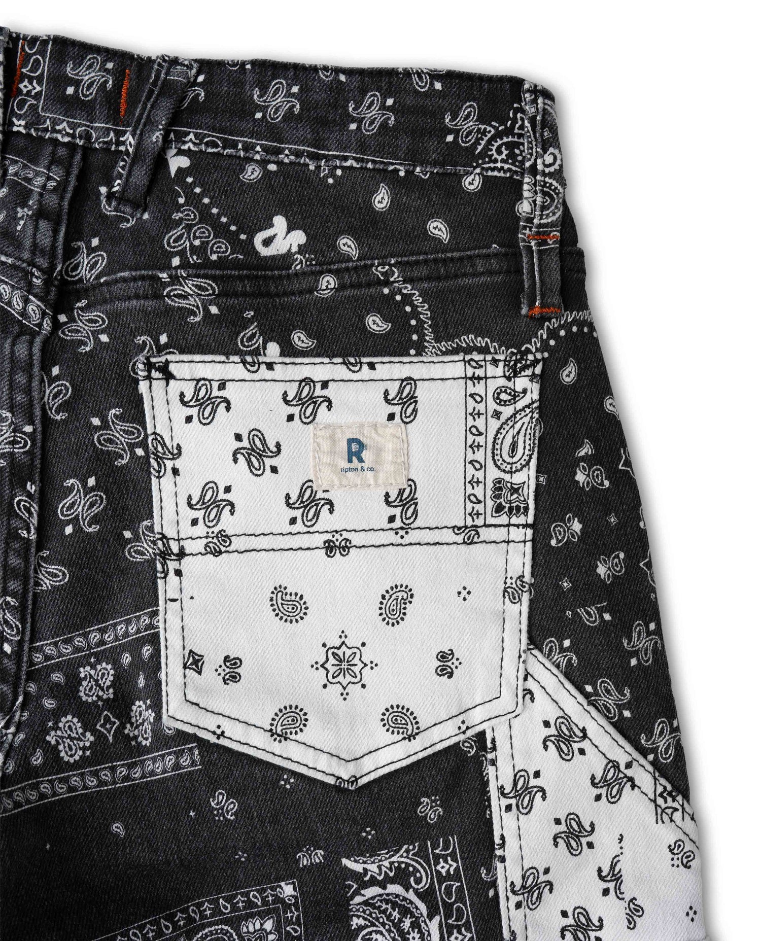 Close-up of the back pocket area of black denim jeans featuring a white bandana paisley print overlay and a white patch pocket with a small blue 'R' logo.