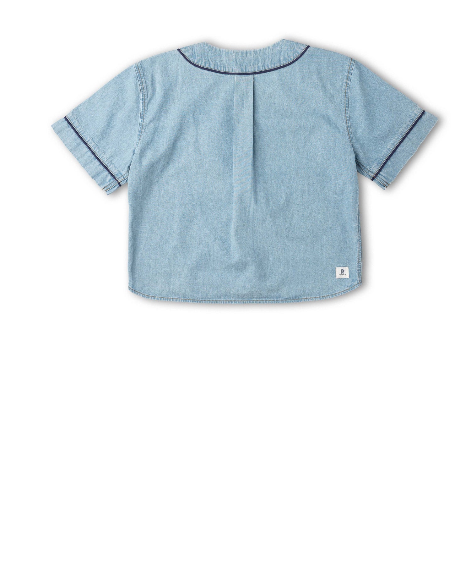 A light blue denim short-sleeve top shown from the back, featuring a central pleat and dark contrast piping along the collar and sleeve cuffs, set against a plain white background.