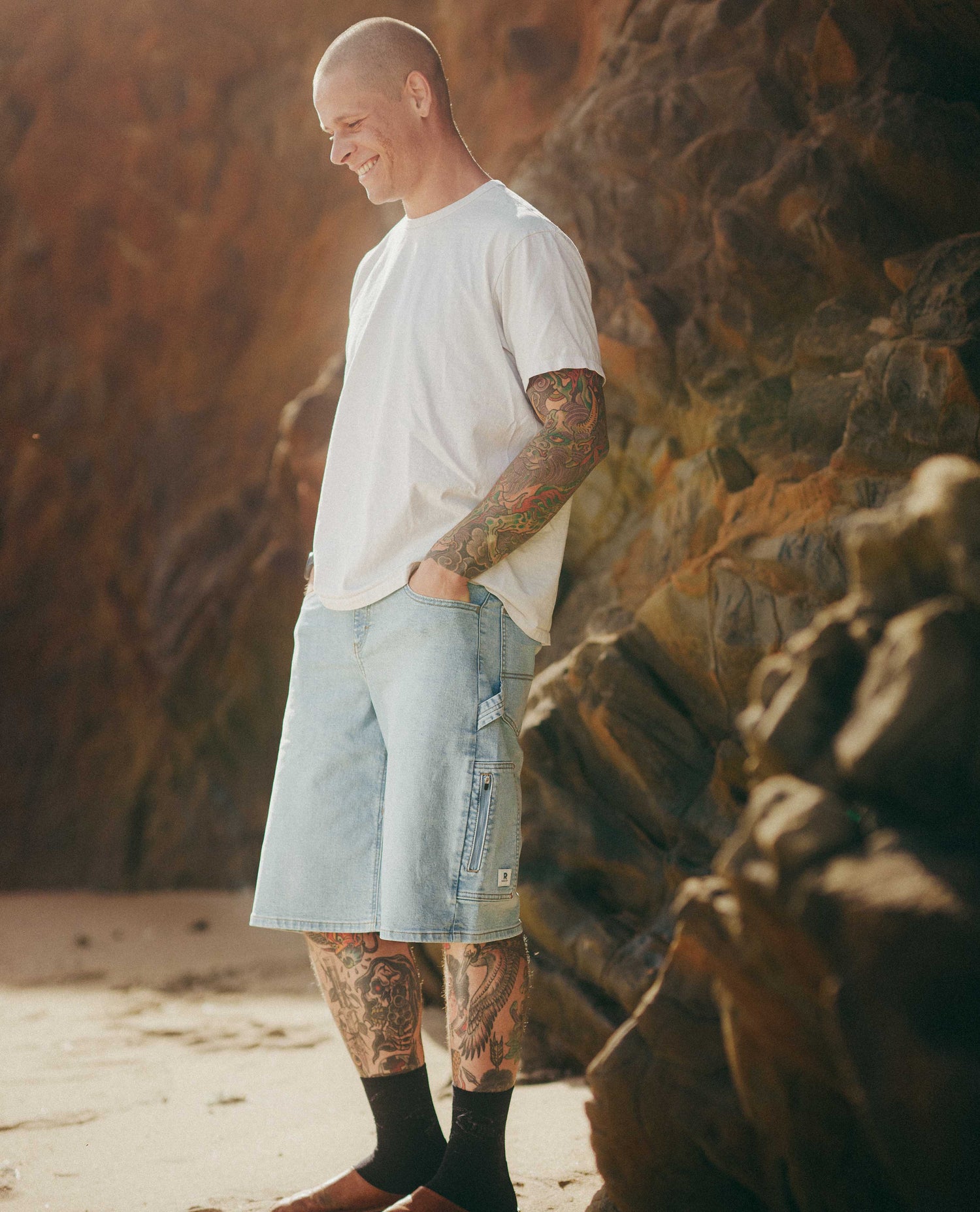 Man with tattoos standing on a beach with rocky cliffs in the background