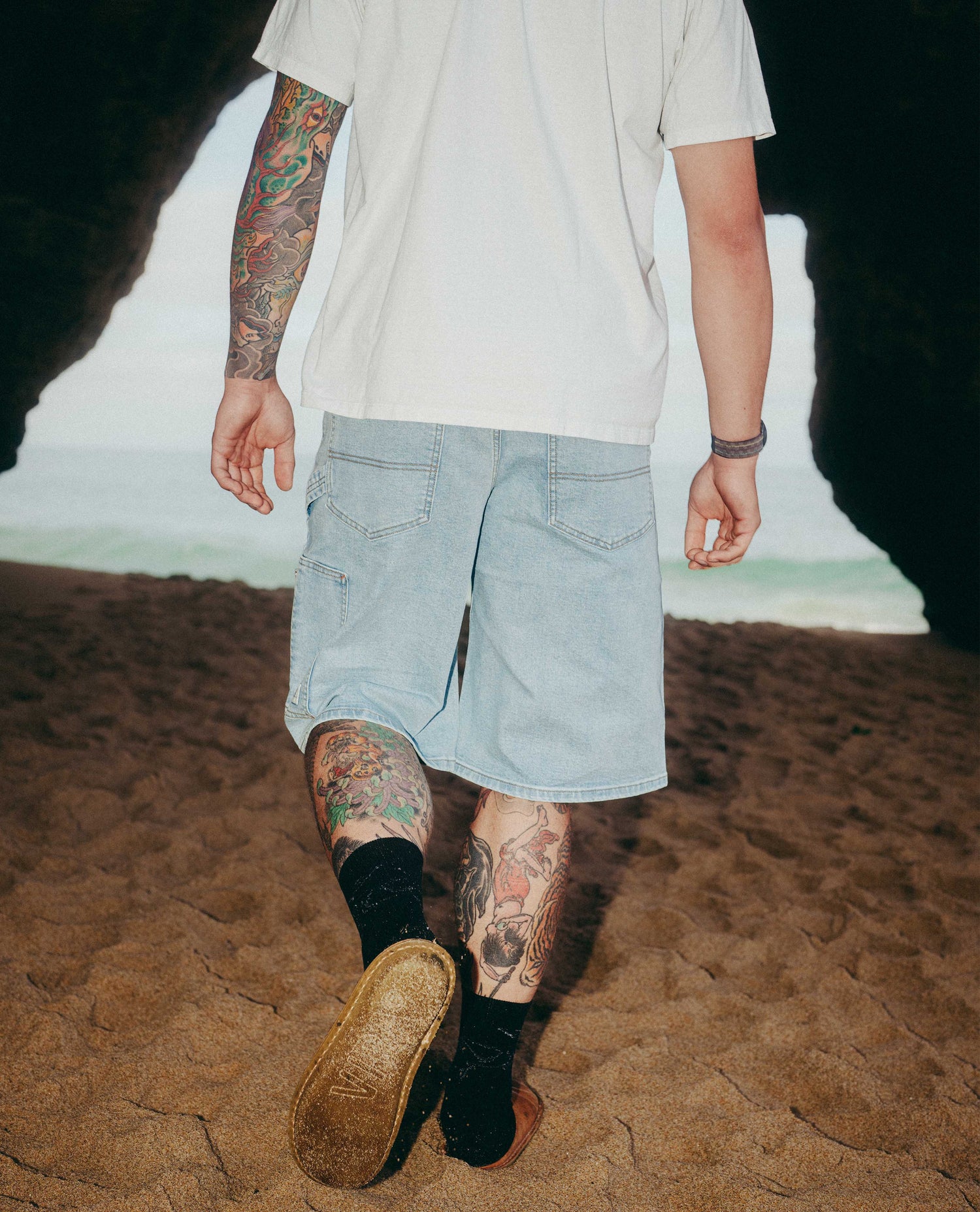 Person wearing light blue shorts and black shoes with tattoos on arms and legs, standing on a sandy surface.