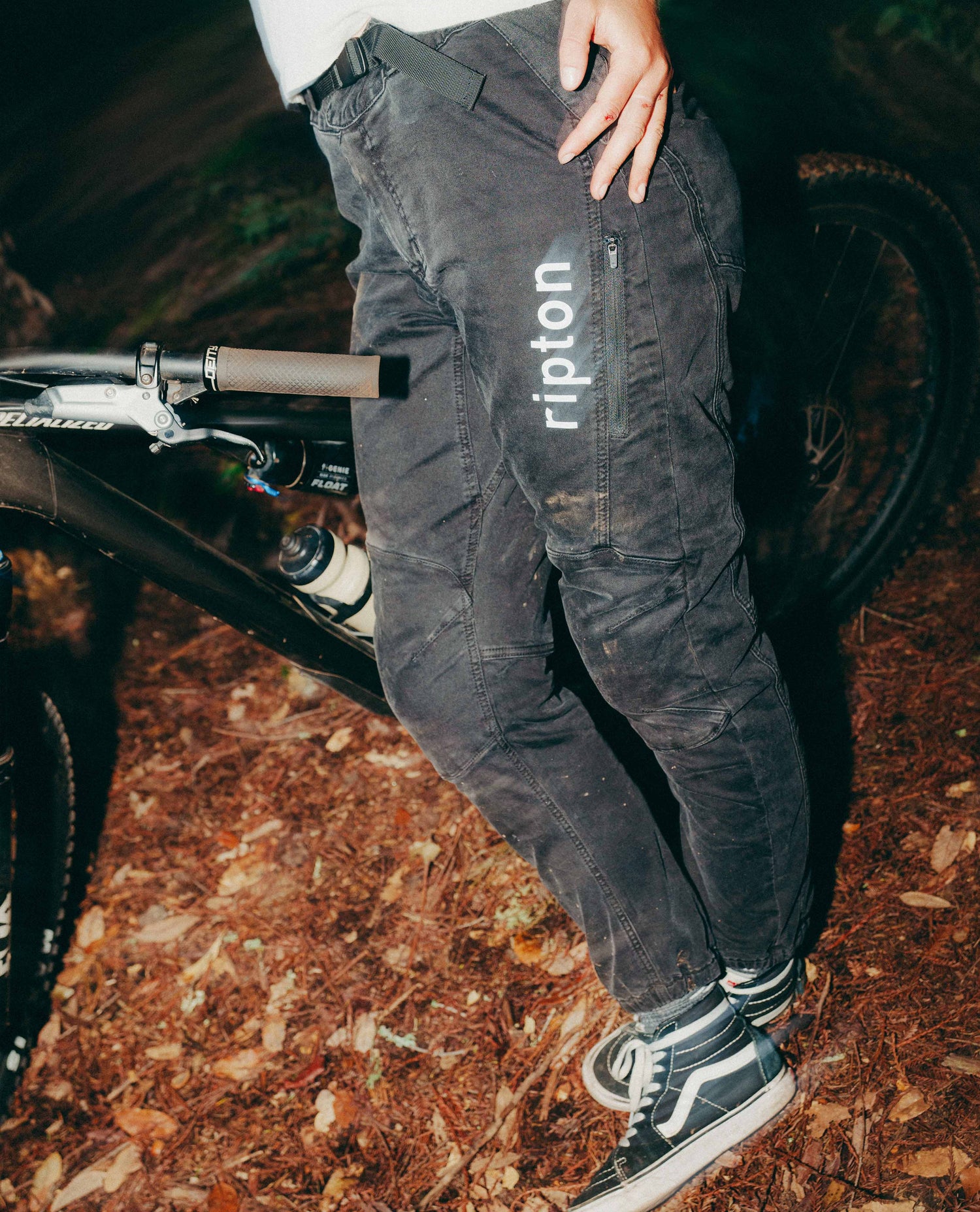 Person wearing black pants with 'ripton' branding, standing next to a bicycle on a trail.