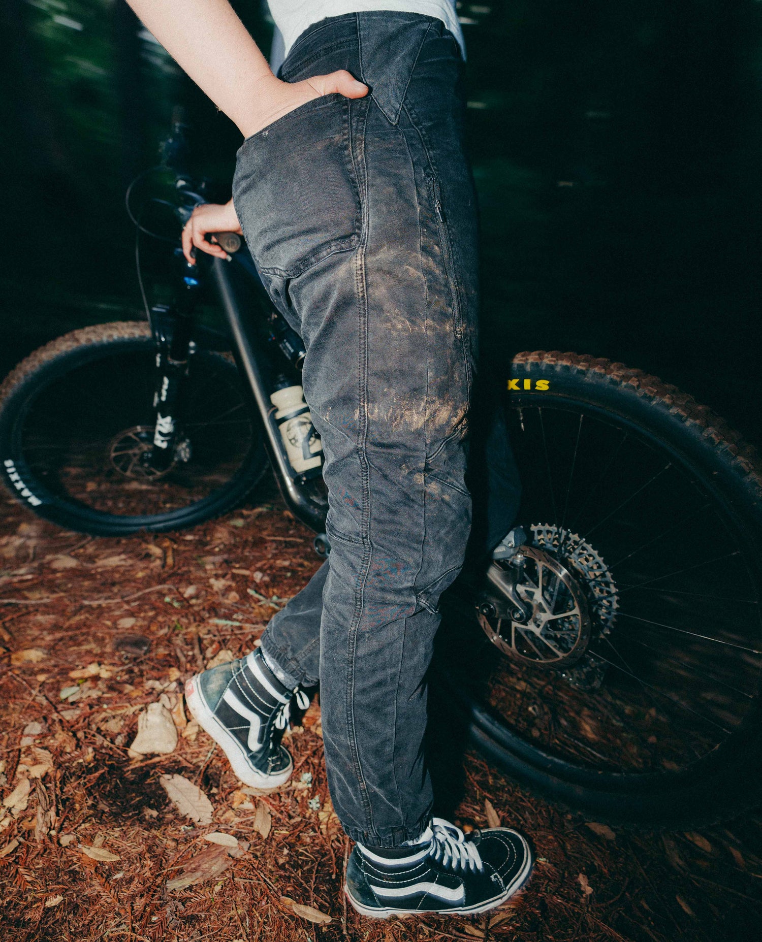 Person wearing dark jeans and sneakers standing next to a bicycle on a forest floor.