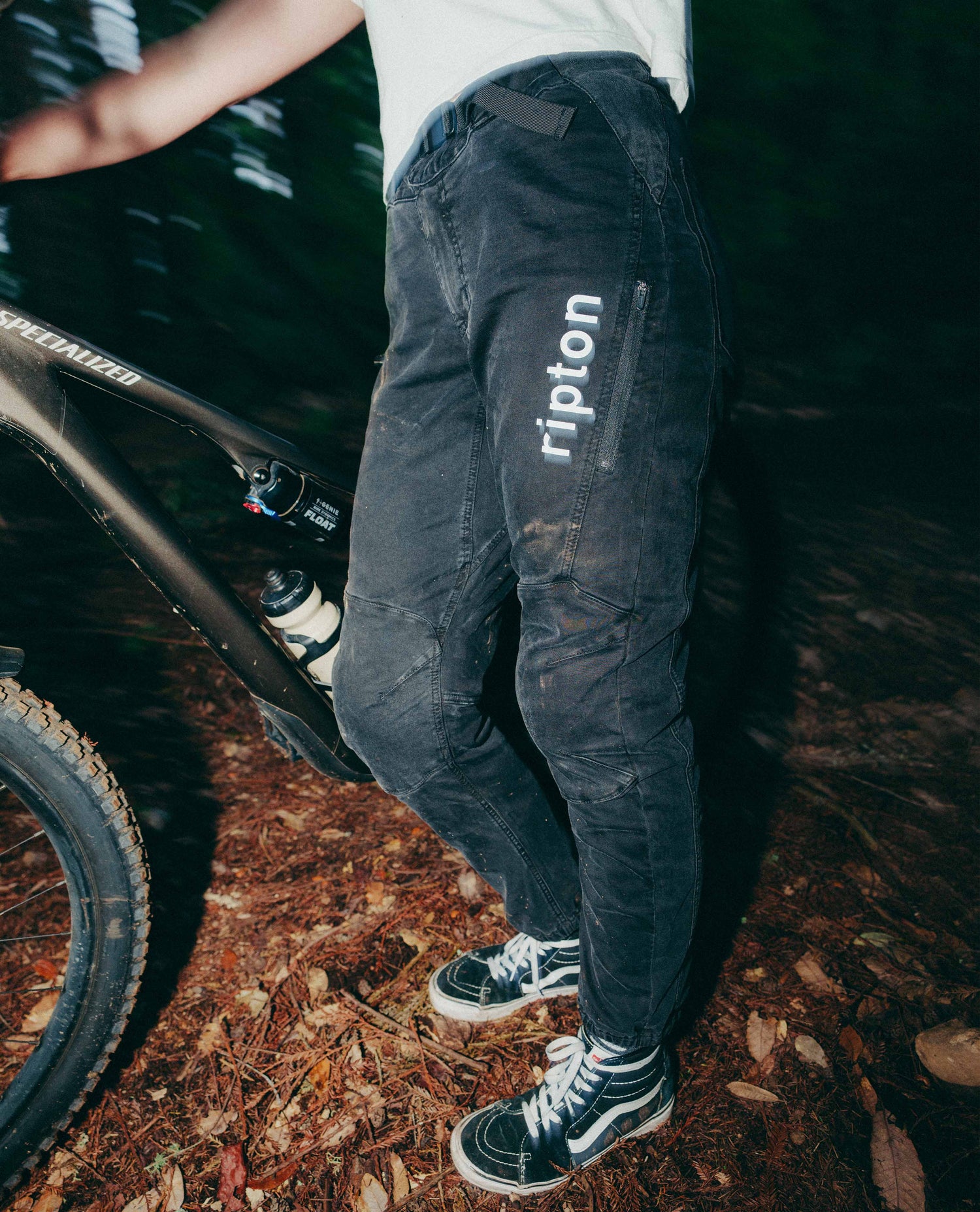 Person wearing dark pants with 'ripton' branding, standing next to a bicycle on a forest floor.
