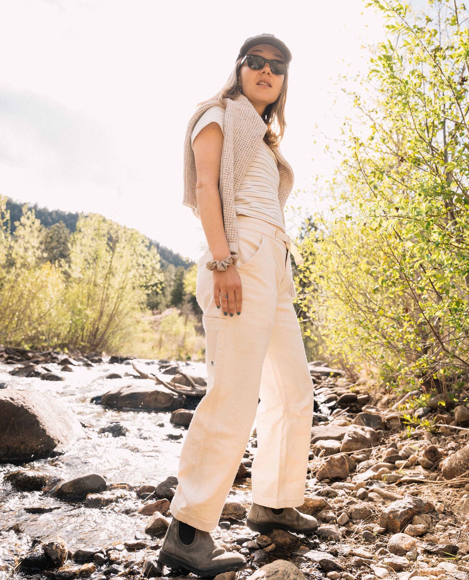 Person wearing a beige outfit standing by a stream with trees in the background