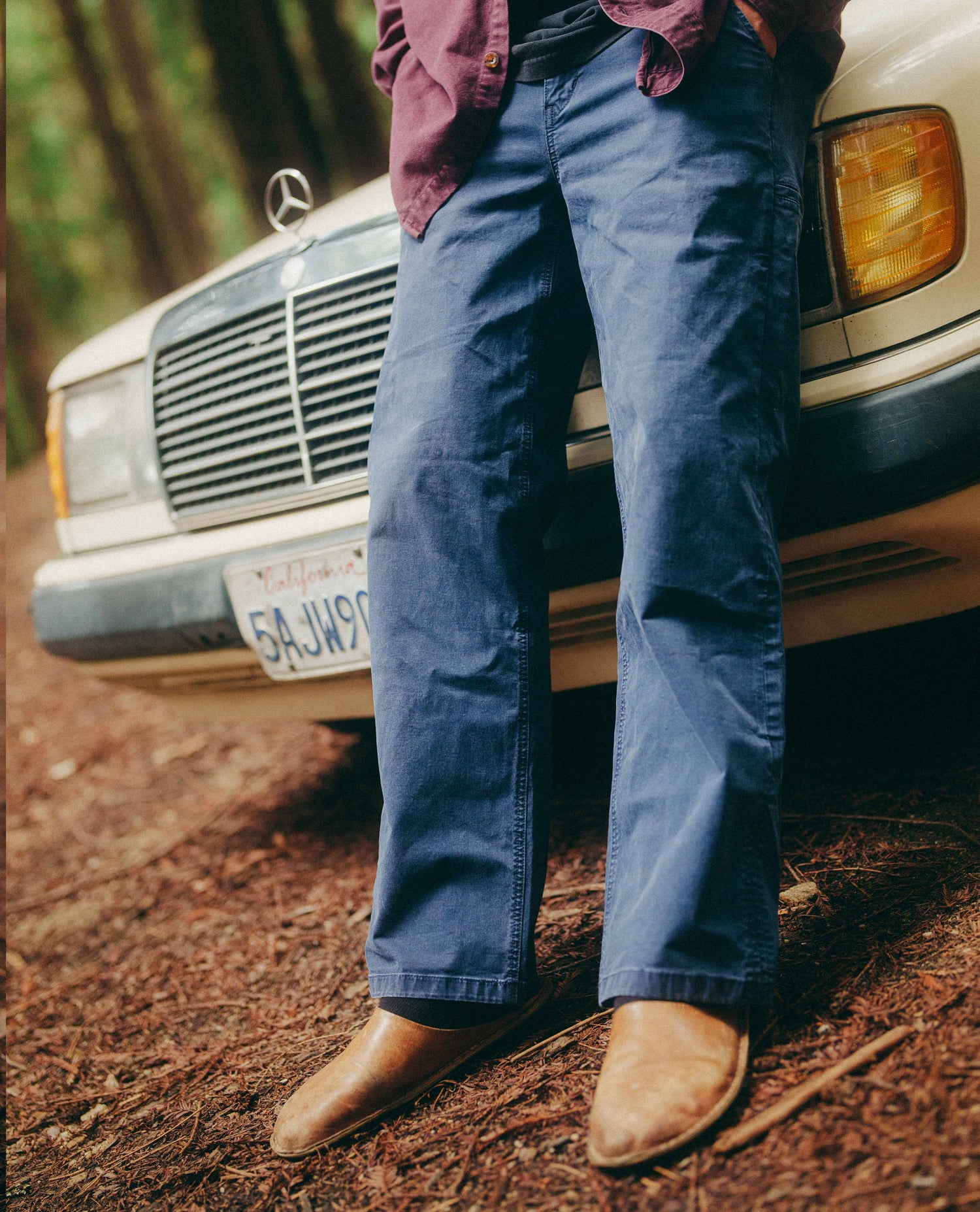 Person wearing blue jeans and brown shoes standing next to a vintage Mercedes-Benz car on a dirt road.