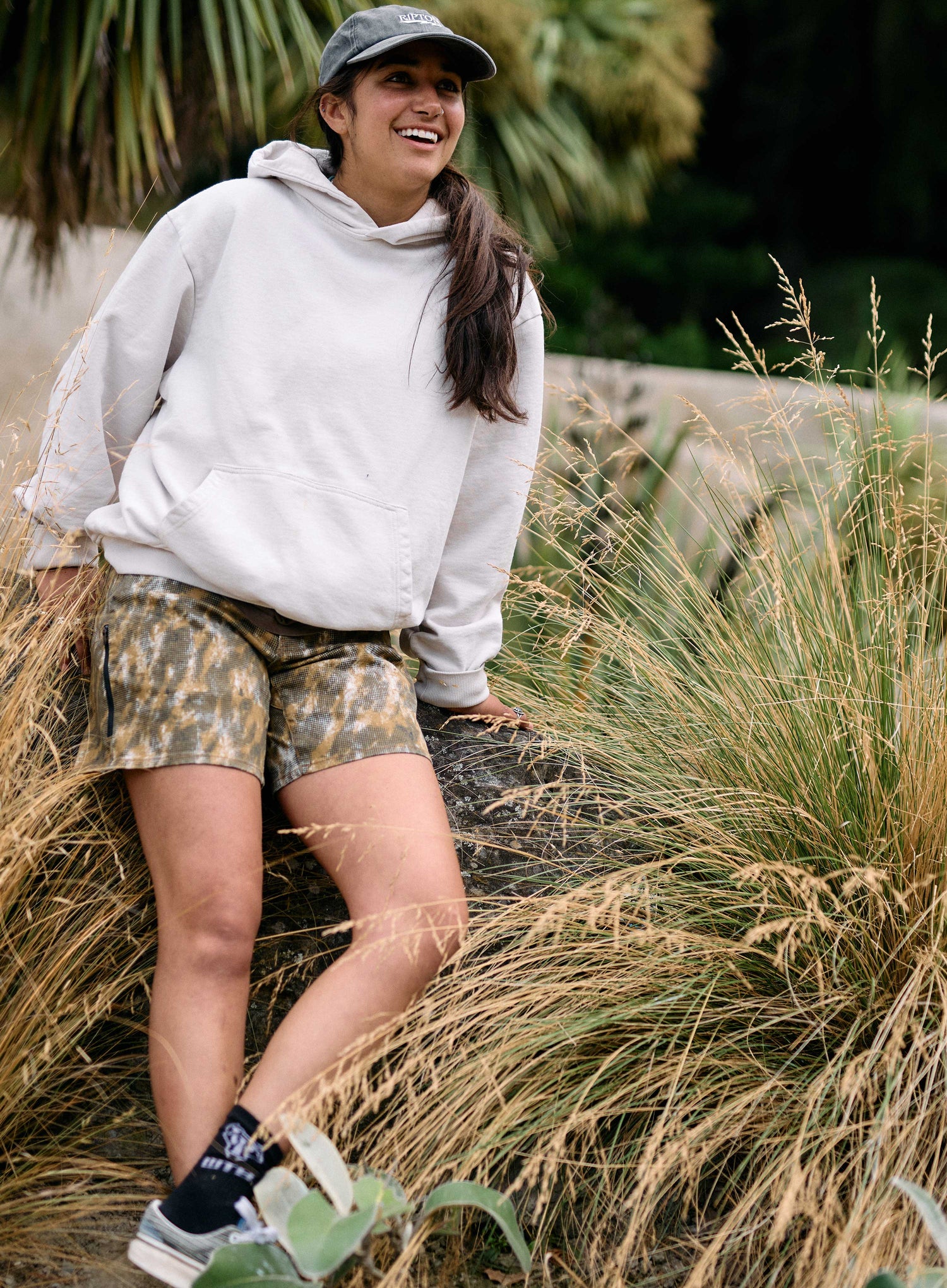 Woman in a gray hoodie and camouflage shorts standing in tall grass.