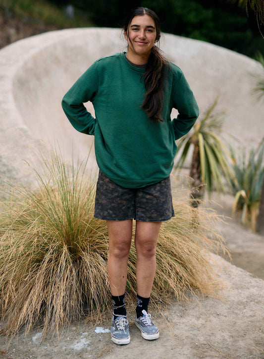 Person wearing a green sweatshirt and camouflage shorts standing in a natural setting with plants and a curved wall.