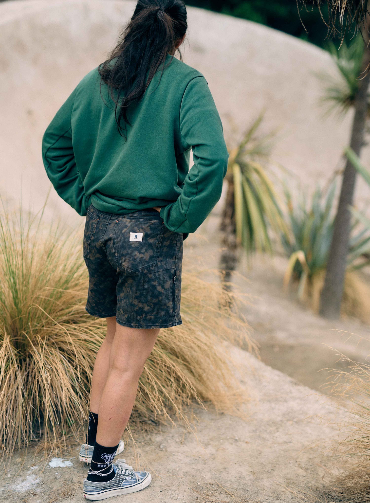 Person wearing a green sweatshirt and camouflage shorts standing in a natural setting with plants and grass.