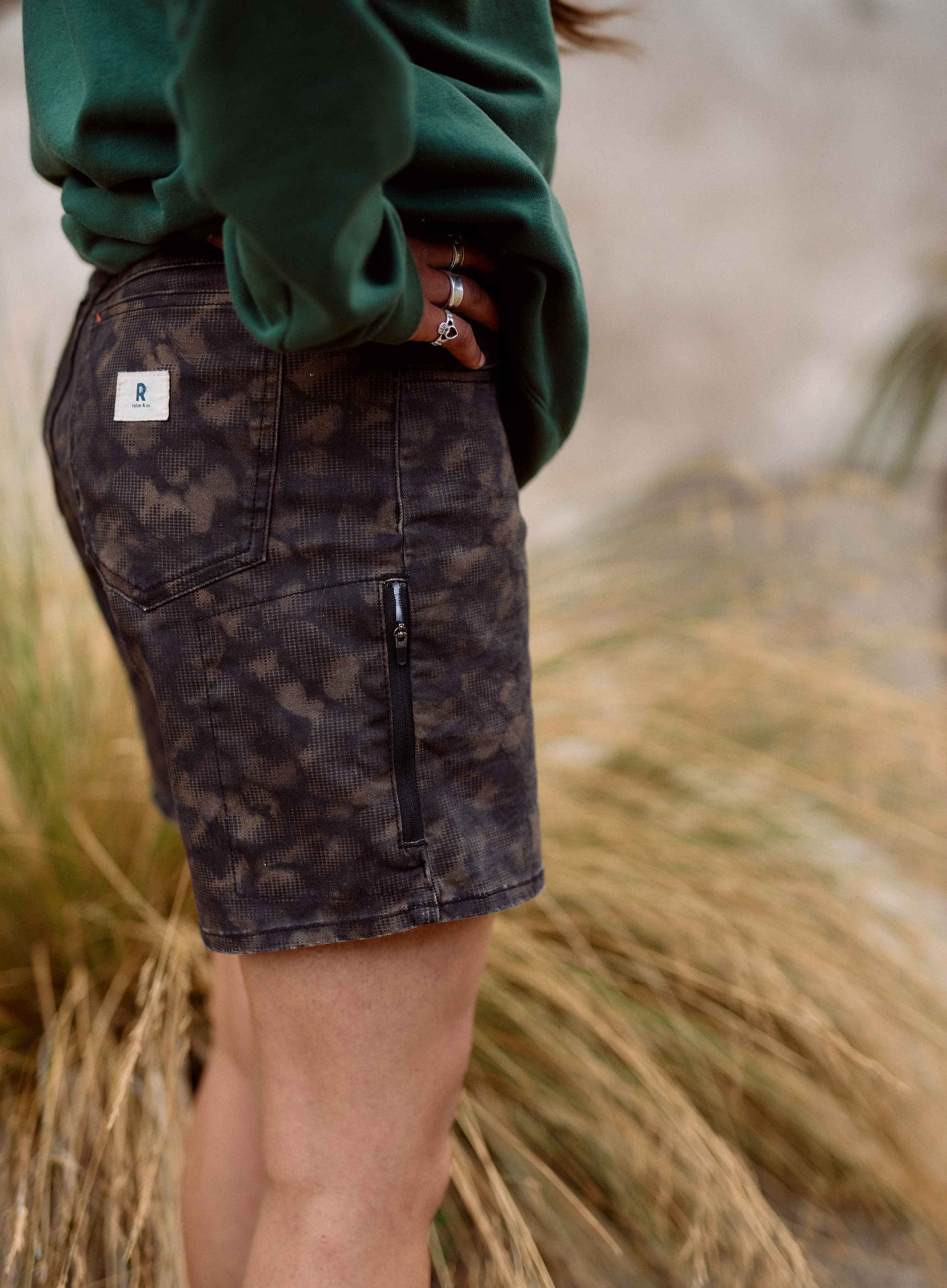 Person wearing a green sweatshirt and camouflage shorts standing in a natural setting with tall grass.
