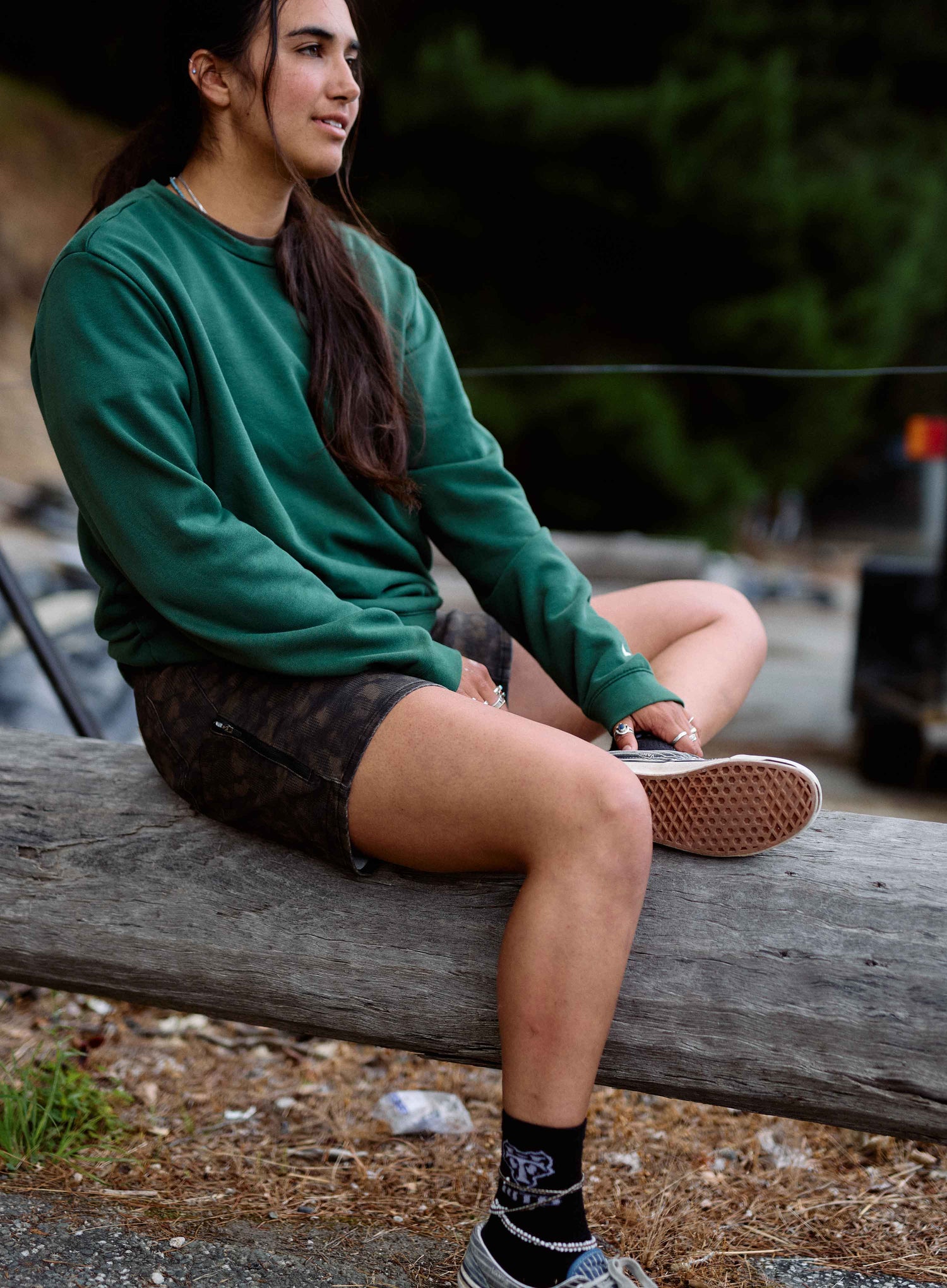 Person sitting on a log outdoors wearing a green sweatshirt and black shorts.