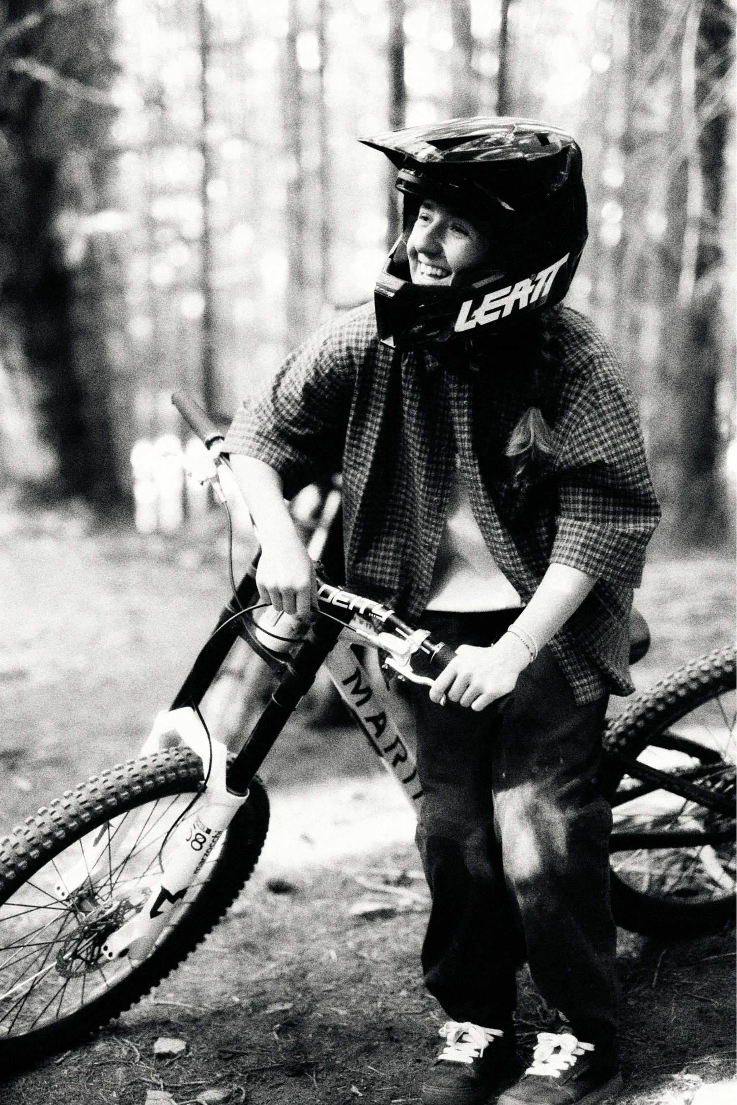 Person with a bicycle in a forest wearing a helmet and plaid shirt.