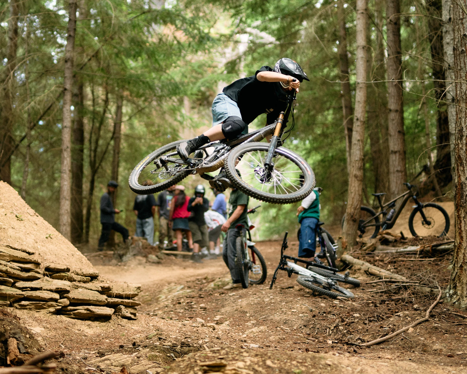 Person riding a mountain bike in a forest setting with other riders and bikes around.