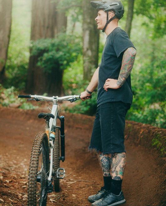 Man with tattoos standing next to a mountain bike on a dirt trail in a forest